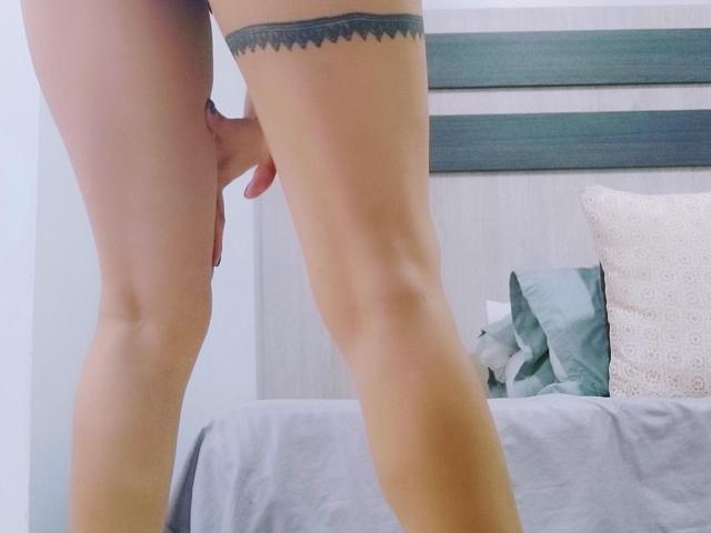 XBellaEvaX's bare legs, standing, with her hand covering her pussy. She has a black lace thigh band, and a bed with pillows in the background.