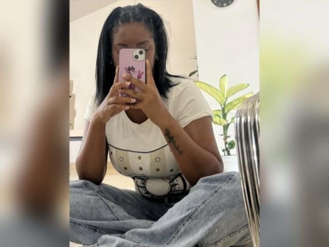 TiffanySammy with dark skin, black pigtails, white t-shirt, and buttoned chest, sitting cross-legged, holding pink phone, showing tattoo on right arm, in mirror selfie.