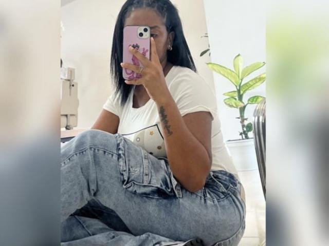 TiffanySammy with black hair, white tee, and blue jeans, sitting cross-legged, holding pink phone, tattoo on right arm, mirror selfie, white room, green plant in background.