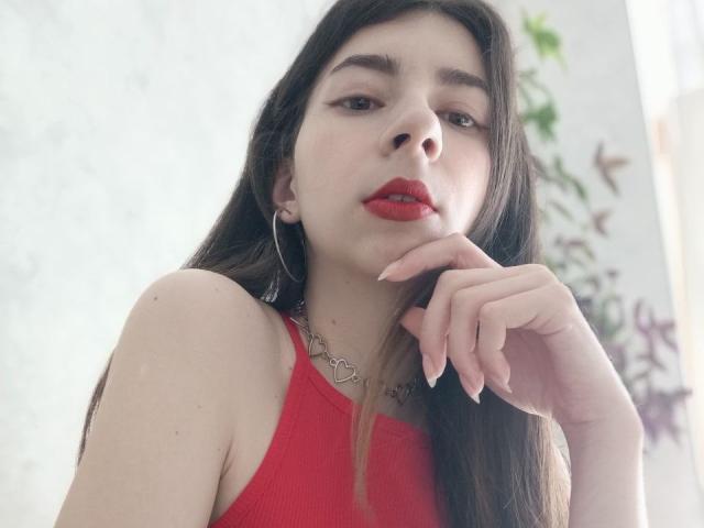 WollyMolly with long brown hair, red lipstick, and red tank top, wearing hoop earrings and a heart necklace, touching her chin, showing off her glossy nails.