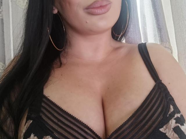 KimStar's big tits in a black lace bra, showing cleavage with her black hair down and gold hoop earrings. Her pink lips are slightly parted.