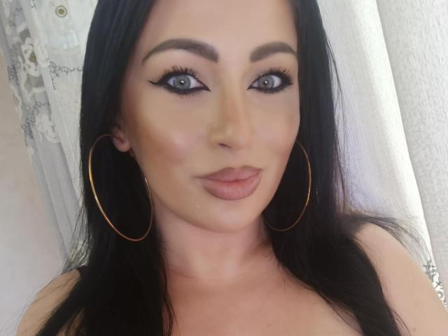 KimStar with black hair, blue eyes, and gold hoop earrings, has full lips and is smiling at the camera, showing off her smooth, tan complexion and winged eyeliner.