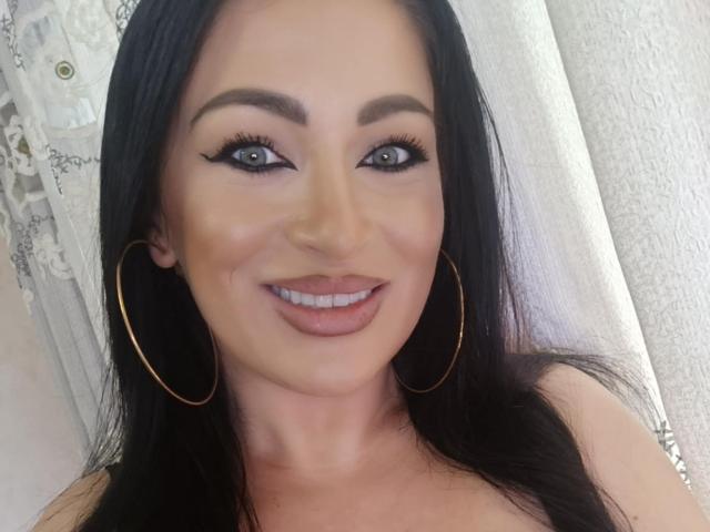 KimStar has blue eyes, dark eyebrows, long black hair, and is smiling with pink lipstick. She's wearing gold hoop earrings and has a cute, sexy face.