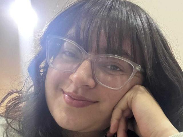 MeganFw with dark wavy hair, wearing clear glasses, has a cute smile and a slightly flushed face, resting her head on her hand, looking relaxed.