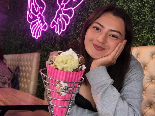 MeganFw with dark brown hair, smiling, holding a pink wire basket with ice cream and chocolate, resting her head on her hand, wearing a gray top, black bra visible.