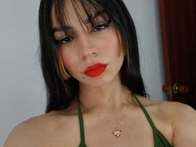 KatherinJohns has long black hair, red lipstick, green halter top, gold necklace with heart pendant, and a small mole on her chin. She has dark eyeliner and a cute face.