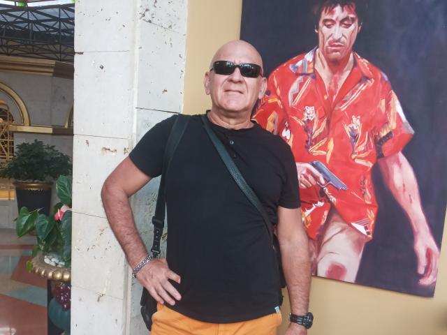 Una coppia di mezza età, calva, con occhiali da sole, una maglietta nera, pantaloni gialli e una borsa verde, è in piedi accanto a un dipinto rosso con motivi floreali raffigurante un uomo che tiene una pistola..