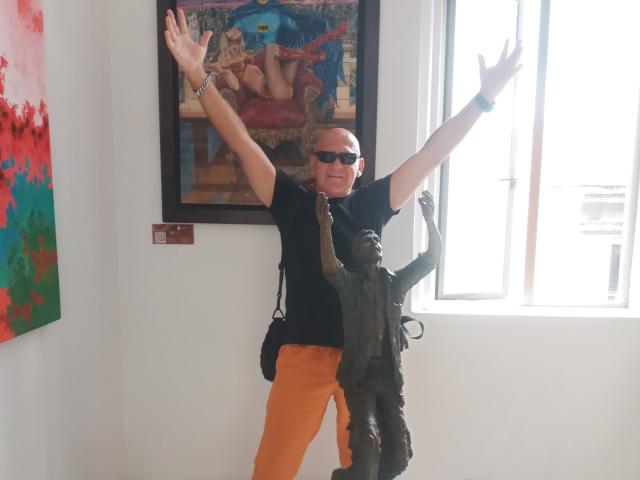 CoupleMature with arms raised, bald, wearing sunglasses, black shirt, orange pants, standing next to a bronze statue of a naked woman. Painting with blue and red colors in background.