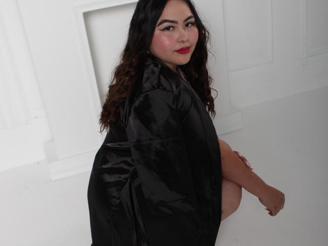 ElinaX with wavy dark hair, red lipstick, and black shiny dress, sitting with her back to the camera, looking over her shoulder, showing her bare ass.