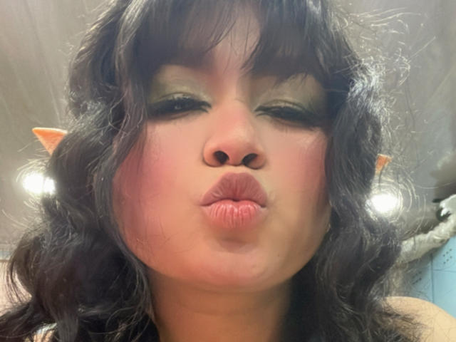 LillyMayer with puffy pink lips, winking, wearing pointy elf ears, and dark curly hair, giving a playful kiss pout. Her green eyeshadow is slightly smudged.