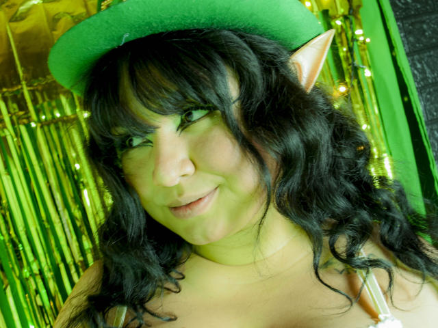 LillyMayer met een groene elfhoed, puntige oren, zwart krullend haar, groene ogen, een lichte glimlach en een groenige huidtint, gekleed in een groene top, en ziet er schattig en aantrekkelijk uit..