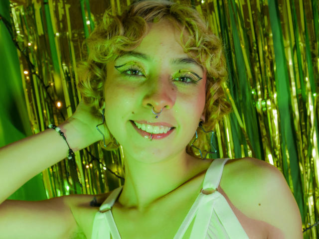 Emily Allen, met krullend blond haar, groene oogmake-up, een neusring en een lip-piercing, lachend, draagt een witte tanktop, heeft groen licht op haar en een armband om, en ziet er blij en schattig uit..