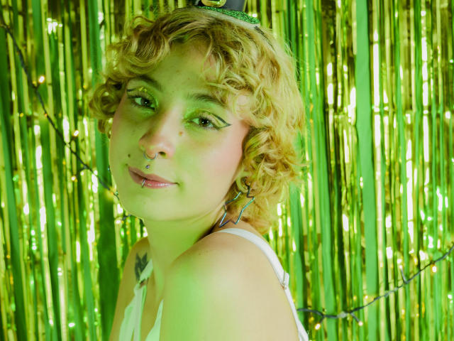 EmilyAllen with curly blonde hair, green eyeshadow, nose ring, and lip piercing, wearing a white tank top, standing in front of green metallic curtains.