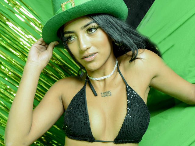 GisellDavies with tan skin, black hair, wearing a green hat, black sequined bikini top, silver choker, and green background, showing small tattoos on chest, looking at camera.