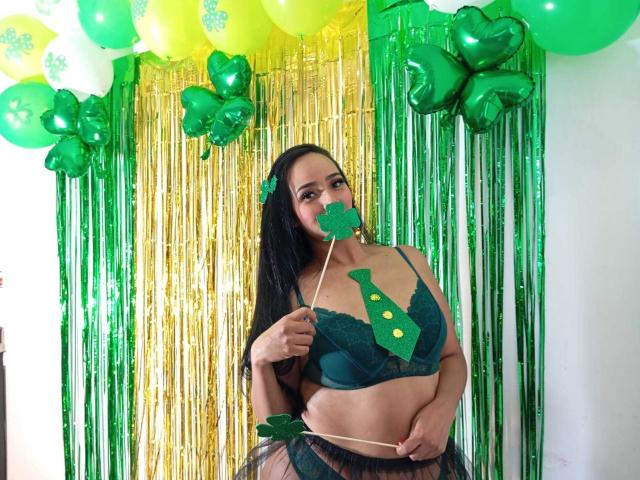 AmberRousell met een groene beha en een schattige groene blad op haar borsten, houdt een groen blad voor haar gezicht, draagt groene en gouden ballonnen en groene linten, in een outfit voor Sint Patrick's Dag..