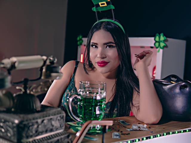 OliviGrace with green headpiece, black hair, red lipstick, green dress, holding drink, table with green glass, cards, and bag, looking at camera, smiling. Boobs visible.
