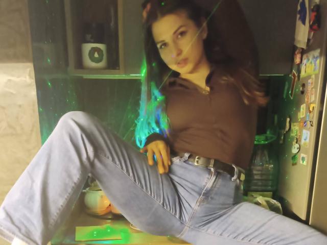 DominikaRos with green glowing hands, brown shirt, blue jeans, and belt, sitting with legs spread, in a dimly lit kitchen, looking at the camera with a relaxed expression.