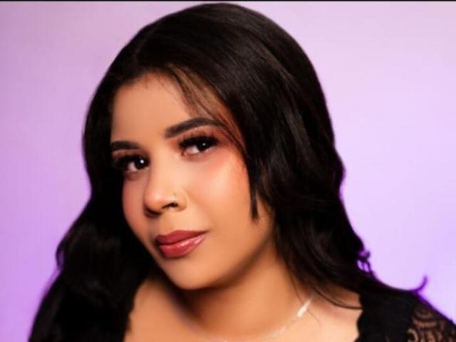 ArianaPrincess a les cheveux noirs et ondulés, des yeux marrons et porte du rouge à lèvres. Elle porte un haut noir et a un sourire subtil et confiant, avec un petit piercing au nez..