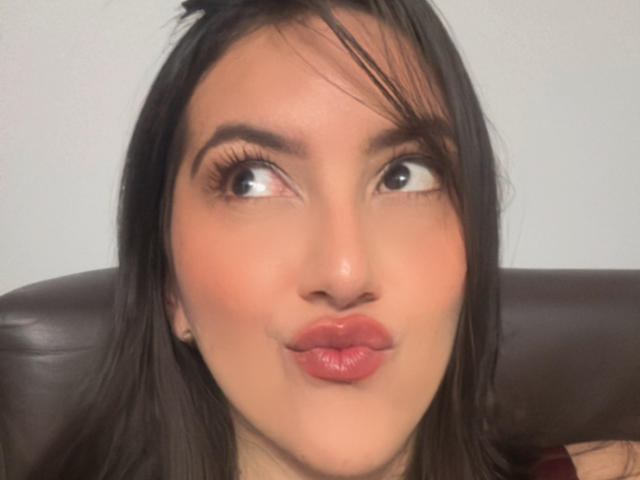 JenaMarquezD with pouty red lips, dark brown hair, and upturned eyes looking to the side, has a fair skin tone and subtle makeup, sitting on a brown leather.