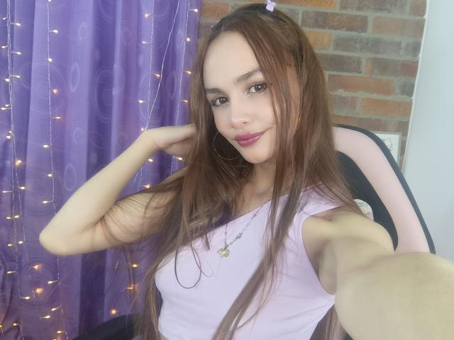 LiliiMoore has long brown hair, pink lipstick, and is wearing a purple tank top with a necklace, taking a selfie with a playful smile. Boobs are small, cute.