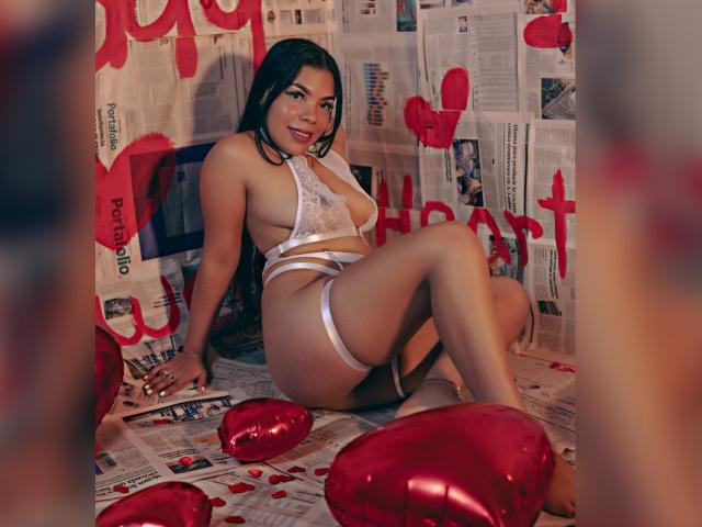 AdrianaPaul with long black hair, wearing white lace lingerie and garter, sitting on newspaper with red heart balloons, showing her ass and white thigh straps.