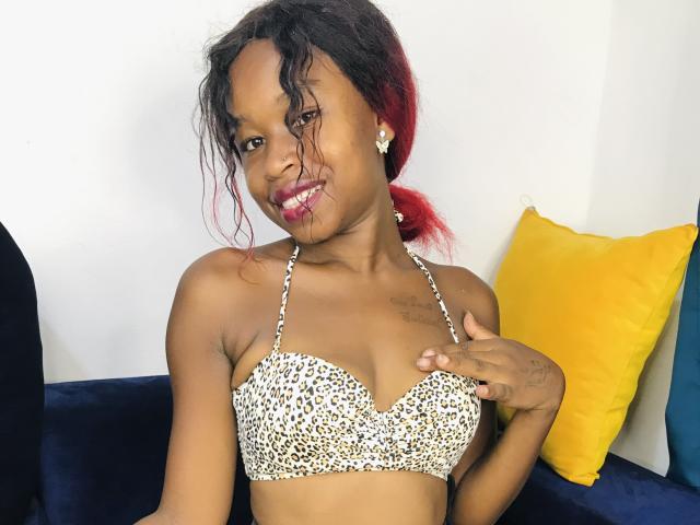 SalmaMagik with dark skin, red-tipped black hair, wearing a leopard print bra, touching her chest, smiling with pink lipstick, yellow and blue pillows in the background.