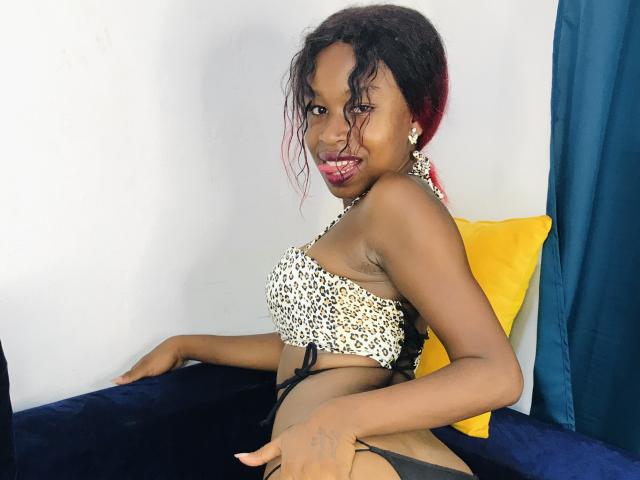 SalmaMagik with dark skin, red-dyed hair, and small breasts, wearing a leopard-print bikini top, black bottoms, and red lipstick, sitting on a blue and yellow couch.