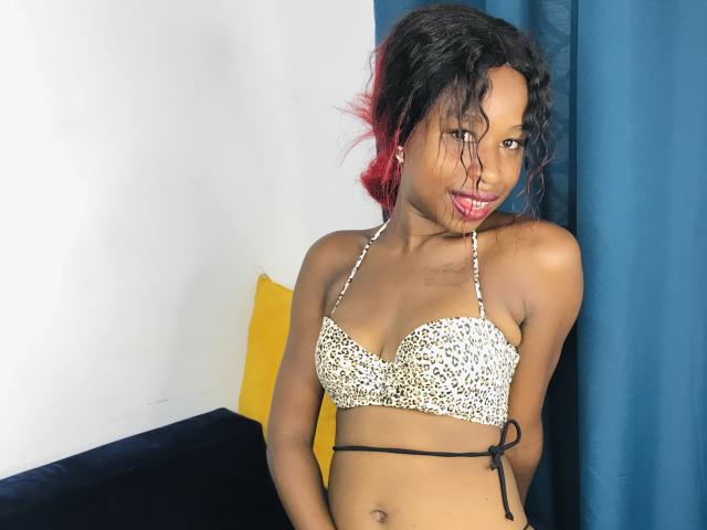 SalmaMagik with dark skin, small boobs in a leopard print bikini top, red-pink hair, smiling, nose ring, blue curtain, white wall, black sofa, yellow pillow, looking at camera.