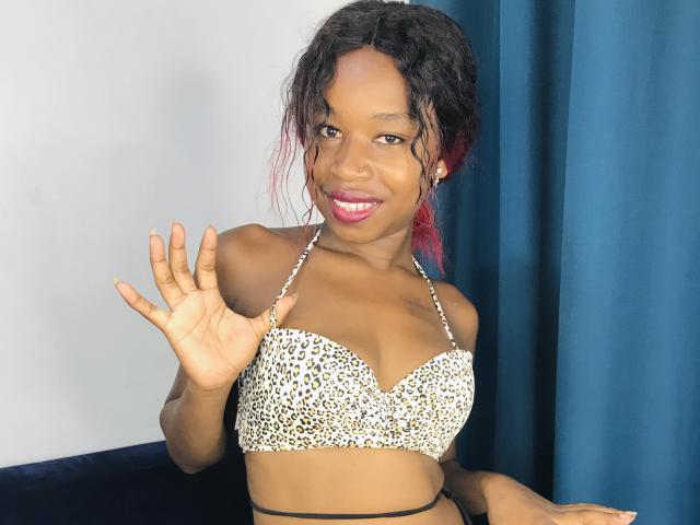 SalmaMagik with dark skin, curly black hair with red tips, wearing a leopard print bra, smiling, waving with her right hand, showing off her small tits.