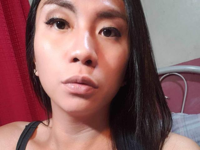 XXSexyCumTsDomxx has shiny brown skin, black straight hair, and is pouting with slightly parted lips. She wears a black tank top and small gold earrings.