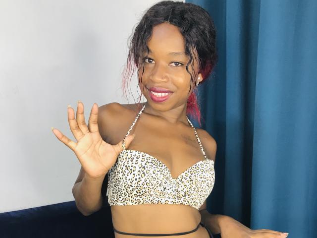 SalmaMagik with dark skin, wavy black hair with red tips, wearing a leopard print bra, smiling, waving with her right hand, showing her tits.