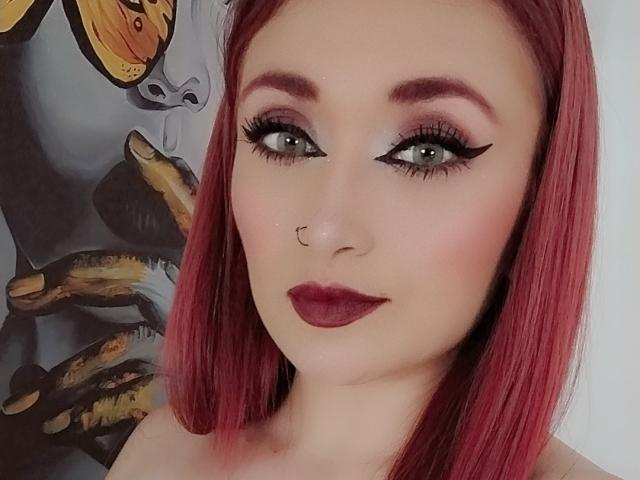 PrincesaLilith has red hair, green eyes with thick black eyeliner, pinkish-gray eyeshadow, dark red lipstick, nose ring, and is looking at the camera with a slight smile.