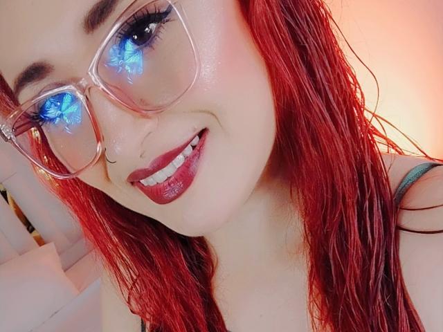 PrincesaLilith with red hair, glasses reflecting blue butterflies, nose ring, and red lipstick, smiling at the camera. Her tits are partially visible, wearing a black strap.