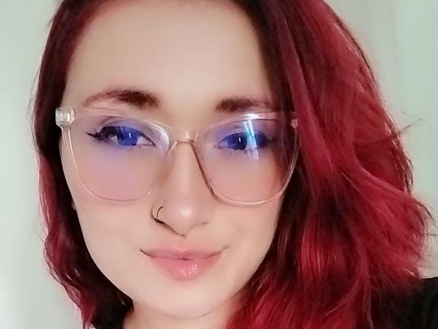PrincesaLilith with red wavy hair, clear glasses, blue eyeshadow, nose ring, and pink lips, smiling softly. Boobs not visible. Ass not visible. Pussy not visible.
