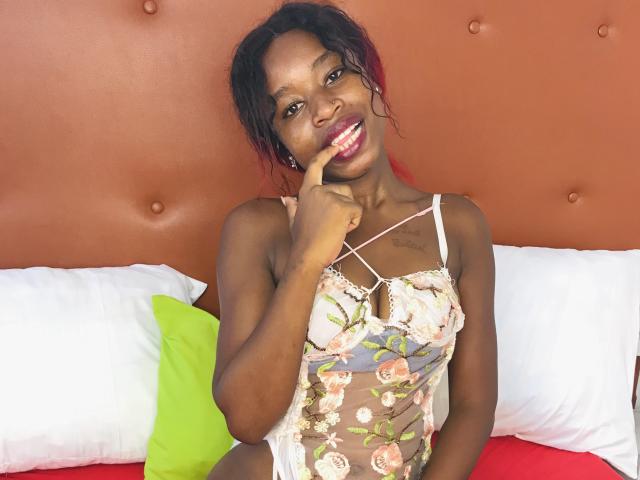 SalmaMagik with dark skin, wearing floral lace lingerie, has her finger on her red-lipped, smiling mouth, showing her small, firm tits and cute, curvy ass.