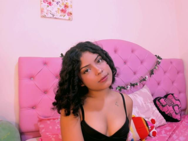 AshleyAmber, avec ses cheveux noirs bouclés, portant un débardeur noir, montrant ses seins, assise sur un lit rose capitonné, regardant la caméra, avec un adorable jouet en peluche..