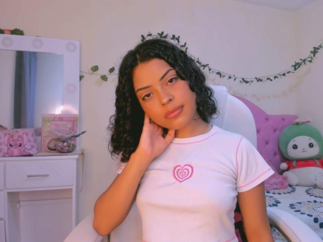 AshleyAmber, avec ses cheveux noirs bouclés, portant un t-shirt blanc avec un cœur rose, assise sur une chaise, une main sur sa joue, regardant la caméra, dans une pièce mignonne aux couleurs roses..