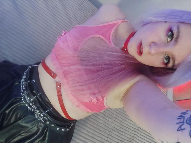 WhiteKrol met roze haar, een rode choker en een roze top, die haar borsten laat zien, draagt zwarte leren broek met een kettingriem, kijkt naar de camera, heeft een tatoeage op haar arm, met paars en rood licht..