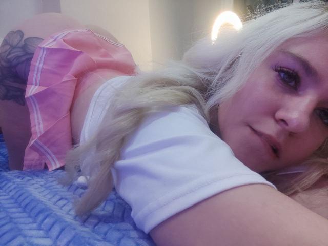 WhiteKrol con capelli biondi, gonna rosa e top bianco, in posizione a quattro zampe, mostra il sedere e il tatuaggio, guardando la telecamera con un leggero sorriso. Sotto di lei, una coperta blu..