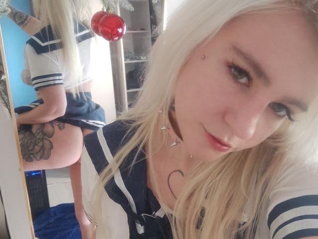 WhiteKrol with blonde hair, blue sailor outfit, tattoo on left thigh, mirror reflection showing her ass, choker necklace, and a piercing below her lip, looking at the camera.