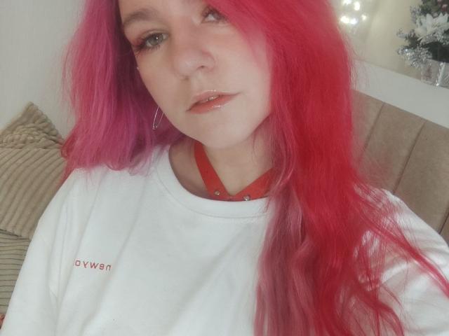 WhiteKrol, con el cabello de un rosa brillante, vestida con una camisa blanca, un collar rojo y aros grandes, tiene labios rosados y brillantes y una expresión neutra, mostrando una apariencia sutil y sexy..