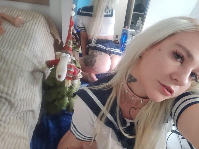 WhiteKrol, con cabello rubio largo, vestida con un traje de marinera azul y blanco, mostrando su trasero en un espejo. Está en una cama con un peluche y una manta azul..