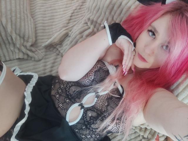 Eine Frau mit weißen Haaren und rosa Strähnen, die ein schwarzes Spitzenoberteil und Armbänder trägt, zeigt ihre Brüste in einem Dienstmädchen-Outfit und liegt auf einer gemusterten Decke..