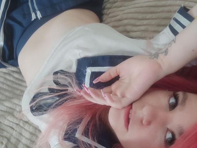WhiteKrol met roze haar, liggend op een bed, gekleed in een marineblauw en wit schoolmeisjeskostuum, met een hand voor haar mond, een zichtbare tatoeage op haar arm en een roze manicure..