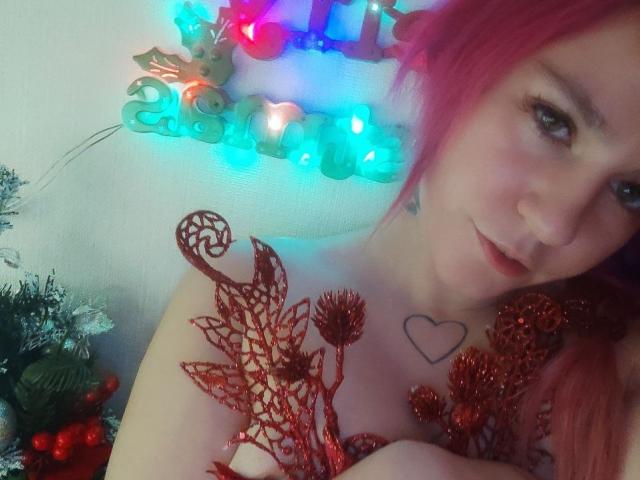 WhiteKrol avec des cheveux roses, un tatouage de cœur sur la poitrine, des décorations en forme de feuilles rouges et scintillantes recouvrant ses seins, des guirlandes de Noël bleues et vertes en arrière-plan, un léger sourire, une lumière douce, un mur blanc..