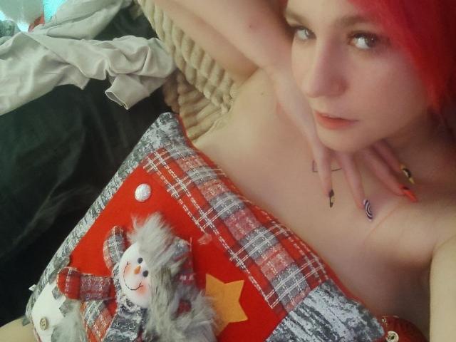 WhiteKrol with red hair, lying down, topless, covering boobs with a red plaid pillow with a fluffy cat and yellow star, hand near face, on a messy bed.