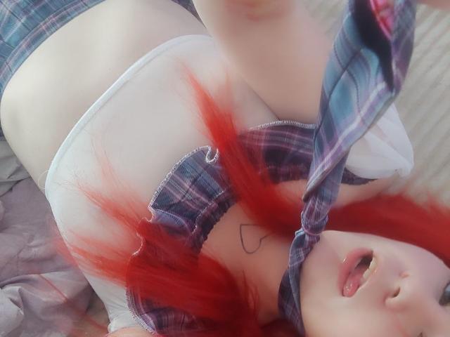WhiteKrol with red hair, lying down, tongue out, wearing a plaid shirt, white bra, and plaid shorts, exposing her small tits, and a heart tattoo on her chest.