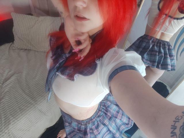 WhiteKrol, com cabelo ruivo vibrante, vestindo uma saia xadrez e um cropped branco, mostrando pequenos seios e uma tatuagem visível no braço direito, em um selfie no espelho..