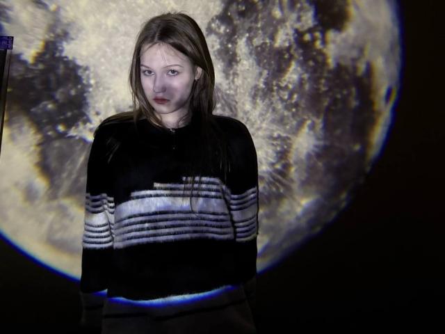Bubochka with pale skin, brown hair, and red lipstick, wearing a black and white striped sweater, standing in front of a large, illuminated, textured moon.