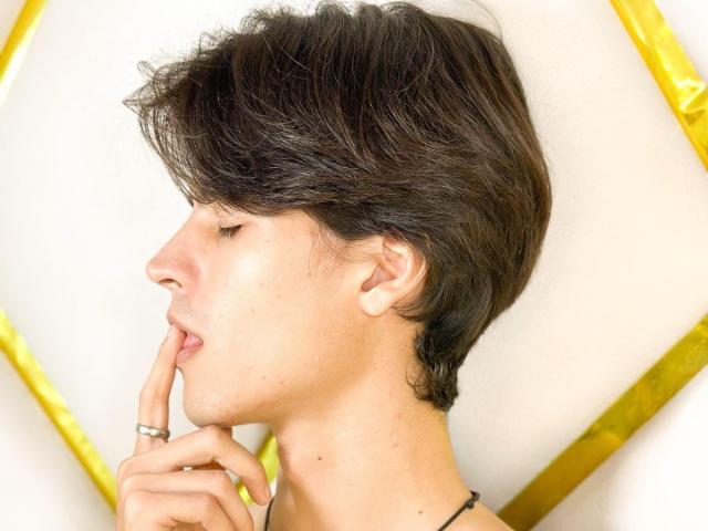 TylerMulber with short, messy brown hair, eyes closed, gently biting finger with a ring on it, light skin, subtle facial stubble, and a black strap visible.